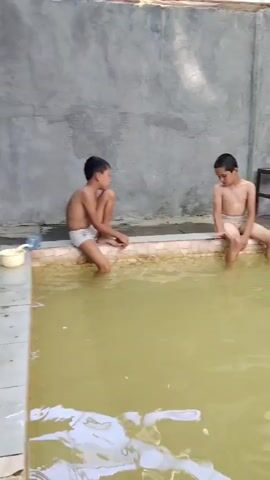 The video opens with a close-up of two young boys sitting on the edge of a pool, with their legs dangling in the murky water. The pool's edge is made of tiles with a patterned border. The wall behind them is a textured grey concrete. The boys are wearing swim shorts. The camera then pans to the right, revealing an older man sitting in the pool, also with his legs submerged. He is wearing black swim trunks. In the background, there's a bench with clothes hanging on a rack behind it. The camera continues to pan, showing a pipe coming out of the wall and a stream of water flowing from it into the pool, creating ripples on the surface. The water appears somewhat discolored, possibly due to minerals or algae. The scene is outdoors, as suggested by the lighting and the open-air structure of the surrounding area. A hollowed-out log or pipe is visible protruding from the water in the foreground. The video ends with a wider shot of the pool and the people in it.