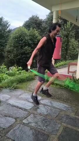 A man is jump roping on a stone pathway in front of a building with lush green foliage and trees in the background. He is wearing a black sleeveless vest, gray shorts, and black athletic shoes. The jump rope appears to be green and is in motion. The weather seems overcast.