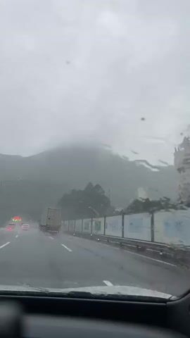 The video, filmed from inside a vehicle, shows a view of a highway during a heavy rainstorm. The windshield is covered in raindrops, distorting the view of the road and the surrounding landscape. In the distance, mountains are visible through the mist and fog. A truck is driving on the highway ahead, and other vehicles can be seen in the distance. A barrier with posters is alongside the highway. The sky is overcast and gray.