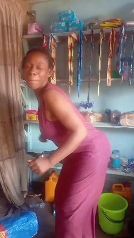 A woman with braided hair is dancing in a small cluttered room. She is wearing a long, sleeveless pink dress. She is bent over with her hands out, moving rhythmically. Behind her is a wooden shelf filled with various items including packages of tissues, wrapped food items, and small containers. To her left is a curtain and below her on the floor is a blue plastic bag and a yellow container. There is also a green bucket visible on the floor. The room appears small and crowded.