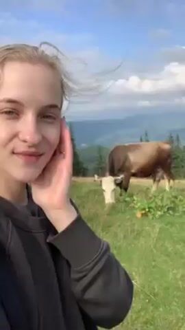 A young blonde woman is recording a selfie video outside in a mountain landscape. She is wearing a black sweater. In the background is a brown and white cow grazing in a green field. The sky is blue with some clouds. There are mountains and trees visible in the distance. The woman has her hand on her cheek and is smiling.