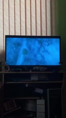 The video shows a television screen displaying a scene with blue-toned imagery and white text that reads "FROM". The screen is positioned on a shelf unit. Above the television, vertical blinds are visible. To the left of the television, on the shelf, is a small circular object. Below the television, on the same shelf, is a dark-colored object, possibly headphones, and a black and white banner with text. The shelf unit itself is dark and appears to have multiple compartments. The lower compartments contain various items, including boxes and what looks like a computer tower on the right side. The overall lighting in the room is dim, with the television screen being the primary light source.