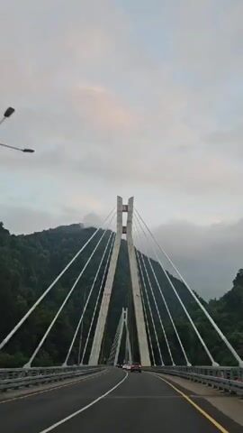 A car drives over a cable-stayed bridge. The bridge has two tall concrete pylons, with cables extending from the pylons to the bridge deck. The bridge is surrounded by lush green mountains. The sky is overcast.