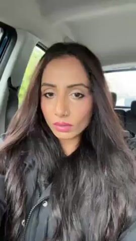 A woman with long, dark hair and pink lipstick is sitting in a car. She is wearing a black jacket. She has a nose ring and is looking directly at the camera. She briefly touches her face with her hand. The background shows the interior of the car and a blurry view outside the window.