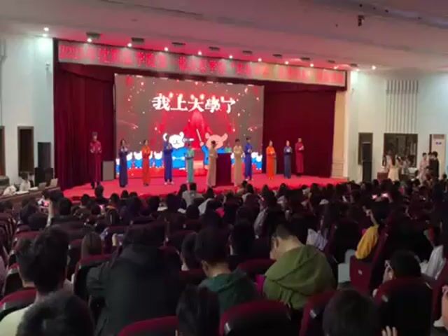 The video shows a large room packed with seated people facing a stage.  On the stage, a group of people, mostly dressed in colorful robes, are performing.  A large screen behind them displays Chinese characters.  The audience appears to be watching the performance and some are recording it on their phones.  The room is well-lit with overhead lighting. The stage is decorated with red curtains.