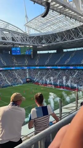 The video shows a wide shot of a soccer stadium from an elevated perspective, looking down at the field. In the foreground, two people, a man and a woman, are standing and watching the game. They are facing away from the camera. The man is wearing a white t-shirt and a tan baseball cap. The woman is wearing a striped shirt with blue and white stripes. The stadium is filled with spectators, many of whom are wearing blue. There is smoke or fog on the field, likely from a fan event or pyrotechnics. The scoreboard is visible in the distance, showing the score as 0-0. The sky is clear and blue, indicating a daytime game. The stadium has a large, modern roof structure.