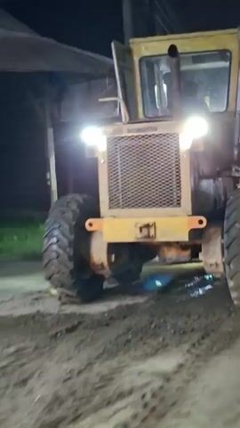 This video appears to be a short, unedited clip taken at night in what seems to be an outdoor area, possibly a street or a yard. The primary subject is a large, yellow construction vehicle, likely a grader or a similar piece of heavy machinery, parked on a dirt or gravel surface. Its headlights are on, illuminating the immediate surroundings. The vehicle is positioned in a way that the front and a large tire are prominently visible.

In the background, there are glimpses of buildings and structures. One building has a sign with text that is partially visible. The sign features logos of various banks and payment services, including "BSI," "ATM," "BCA," "Mandiri," "BNI," "OVO," and "DANA." This suggests the location might be near a business establishment or a public service point. There is also a dark-colored car in the background, with its taillights illuminated, indicating it might be moving or parked.

The overall scene is dimly lit, characteristic of nighttime. There are some reflections and light glares, possibly from streetlights or the vehicle's own lights. The ground appears wet in some areas, suggesting recent rain or damp conditions. No people are clearly visible in the video. The atmosphere is functional, focusing on the machinery and the surrounding commercial environment. The visible text provides specific context about the presence of financial services in the area, which is identified as Blangpeuria, Indonesia.