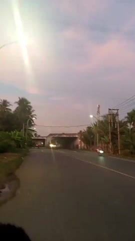 The video shows a road with palm trees on the left and right sides. The sky is a mix of pink and purple hues, indicating either sunrise or sunset. There are streetlights visible along the road. In the distance, there is an overpass or bridge. A motorcycle carrying two people and a large dark object, possibly a musical instrument case, passes by. The motorcycle is moving away from the camera and is illuminated by its headlights. The video appears to be filmed from another moving vehicle. The overall scene is somewhat blurry, suggesting motion.