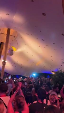 A crowded outdoor event is depicted, with a large, white, tent-like structure overhead illuminated by colored lights, creating a warm orange and pink glow. The ceiling is dotted with dark circular shapes. Numerous people are visible in the foreground, many holding up their phones to record or take pictures. The crowd appears to be enjoying themselves, with some individuals raising their hands. In the center of the frame, a person is being lifted above the crowd, holding up what appears to be a bottle. The lighting shifts to a darker tone towards the end of the clip, with a blurred effect suggesting movement. The atmosphere is that of a lively gathering or festival.