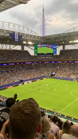 The video shows a wide shot of a soccer stadium filled with spectators. A soccer match is in progress on the green field below, with players in black and white uniforms visible. A large screen above the field displays the game and shows "FIFA WORLD CUP 2026". The stands are packed with a diverse crowd, many of whom are wearing white shirts. The stadium has a modern architectural design with a prominent spire-like structure extending upwards. The sky is overcast, and the lighting suggests it is daytime or early evening. The camera is positioned behind a few spectators in the foreground, whose heads and shoulders are partially visible.