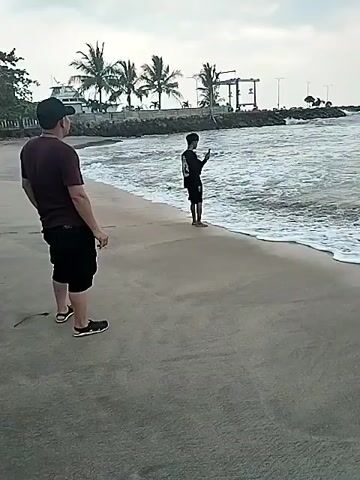 Two people are visible on a beach in front of the ocean. One person is standing on the sand, facing away from the camera, wearing a dark shirt and shorts, and a baseball cap. Another person is standing in the shallow water, facing the camera, wearing dark clothing. In the distance, there are palm trees and structures along the coastline. The ocean has moderate waves and foam. The sky is overcast.