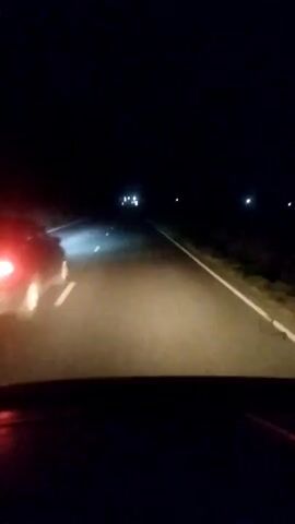 This media file shows a first-person perspective from inside a vehicle driving at night. The road ahead is illuminated by the headlights of the vehicle and the taillights of a car in front, which has its brake lights illuminated, suggesting it is slowing down or stopping. The scene is dark, indicating it is nighttime. The road appears to be a rural highway with a white line marking the lane. There are some faint lights visible in the distance, possibly from other vehicles or buildings. The overall atmosphere is that of a night drive, with a sense of movement and the challenges of low visibility. The location context provided is Akhali Dioknisi, Georgia. There is no visible text in the media file.