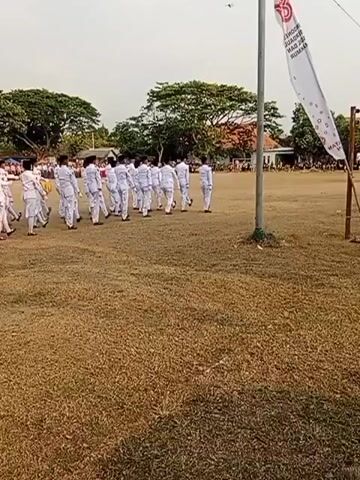 A group of people in white uniforms are marching in formation on a grassy field. They are moving from left to right in the frame. A flagpole with a white banner is to the right of the frame. The banner has red text and a red logo. The people are all facing away from the camera. There are trees and buildings in the background. The sky is overcast.
