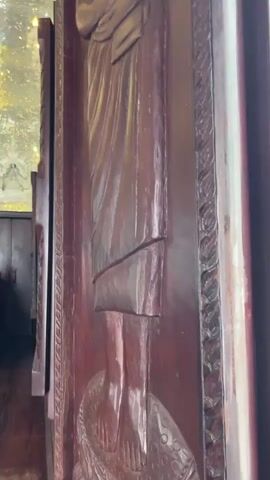 The video shows a view through a partly opened wooden door, revealing the interior of a temple. The door has a carved figure of a person on it. Inside, a large silver Buddha statue sits on a tiered platform, surrounded by smaller statues. The ceiling is covered in reflective material, creating a mirrored effect, and several ornate chandeliers hang from it. The room has dark wooden accents and what appears to be a dark-stained wooden floor. Some flowers are placed around the base of the Buddha statue.