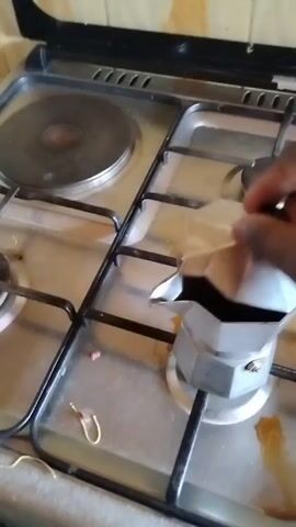 The video shows a Moka pot on a gas stovetop. The Moka pot is silver and partially full of coffee, with the lid open. Steam rises from the coffee. The stovetop is stainless steel with black grates. To the left of the Moka pot is an electric burner. There is a small gold-colored object on the stovetop near the bottom left corner. There is also a brown stain on the stovetop to the right of the Moka pot. The background includes yellow and beige tiling. The camera zooms in slightly on the coffee in the Moka pot.
