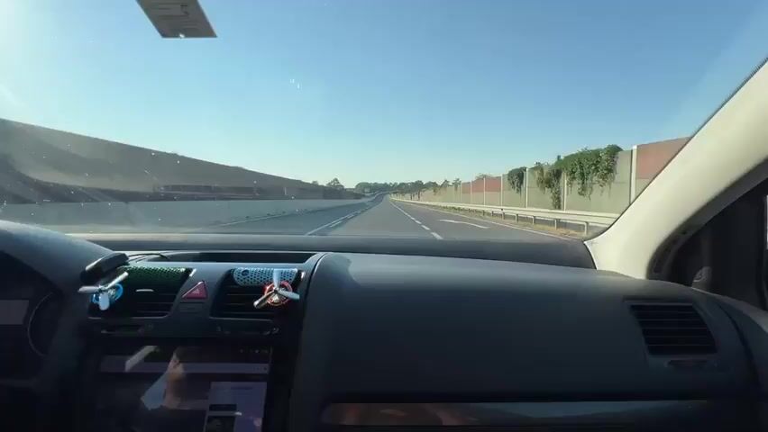 A first-person perspective video shows a person driving a car on a highway. The driver's hands are visible on the steering wheel, and the dashboard is in view, with a navigation screen displaying content. The road ahead is clear, with sound barriers on either side. The sun is shining, creating a bright sky and reflecting off the windshield. The car appears to be moving at a steady pace.