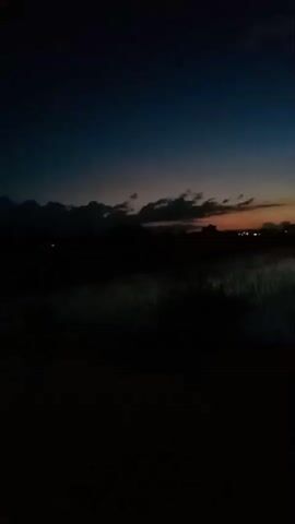 The video captures a serene twilight scene in Bungur, Indonesia, featuring a panoramic view of a rural landscape. The sky transitions from a deep indigo at the top to hues of orange and dark blue near the horizon, indicative of either sunset or dawn. Wispy, dark clouds are visible against the colorful sky on the left side.

In the foreground, the land is dark, suggesting fields or rice paddies, with some faint light reflecting off what appears to be water or dew, creating linear patterns on the ground. As the camera slowly pans to the right, more of these fields come into view. In the distance, beyond the dark land, the scattered, bright lights of what seems to be a town or village are visible, illuminating the distant silhouette of buildings or trees. The overall mood is quiet and peaceful. There are no people or discernible objects in the immediate foreground.