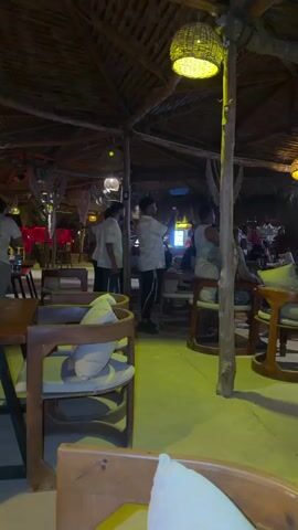 This video shows the interior of a dimly lit, open-air restaurant with a thatched roof. The walls are open to the outside. Several people are seated at tables and chairs, some appear to be staff members in white shirts. There are pendant lights with woven shades hanging from the ceiling. The floor is concrete and appears to be yellow-tinted. In the background, a large screen is visible, displaying a bright image, possibly a television. The atmosphere suggests a casual, possibly beachside establishment.