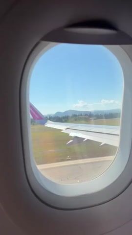 The video shows a view from an airplane window. An airplane wing with a pink and white tail fin is visible. The airplane is on a runway, and the ground outside is a grassy field with some trees and mountains in the distance. The sky is clear and blue. The video is shot in portrait orientation and lasts for a few seconds.