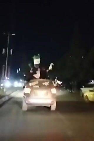At night, a silver vehicle is driving on a street lined with trees and lit by streetlights. Several people stand on the vehicle, waving white flags, and text is written on the rear window. Other vehicles are visible on the street.