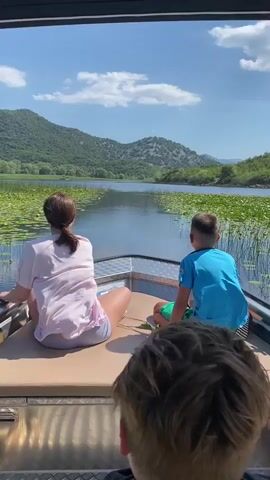 A group of people are on a boat on a lake. The boat is moving slowly. The water is calm and there are lily pads on the surface. In the background, there are green hills and trees. The sky is blue with some white clouds. One person is sitting on the left side of the boat, facing away from the camera. They are wearing a pink shirt and gray shorts. Another person is sitting on the right side of the boat, facing away from the camera. They are wearing a blue shirt and green shorts. In the foreground, the back of another person's head is visible, with blond hair. They are sitting in the boat, facing forward. The boat appears to be a small motorboat.