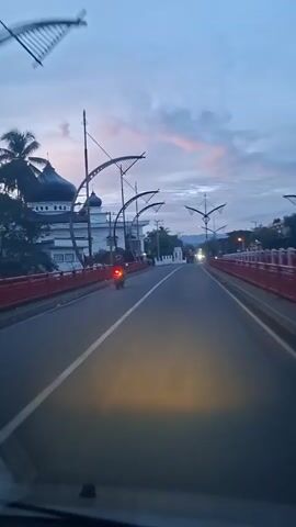 A video from a moving vehicle captures a roadside scene at dusk. On the left, a mosque with a dark dome and white walls is visible. A red barrier runs along the road next to the mosque. The road, which has a white solid line separating two lanes, stretches forward, with the vehicle's headlights illuminating the asphalt. Overhead, strings of decorative lights arch across the road. A motorcyclist with a red taillight drives ahead. On the right, white buildings and trees line the road. The sky is a pale blue with hints of pink and orange near the horizon.