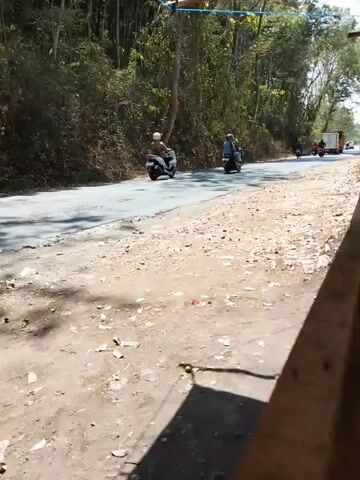 The video captures a road with a shoulder covered in dirt and debris. Several motorcycles are seen riding on the asphalt road. On the left side of the road, there's a steep, wooded incline. On the right side, there's a wooden structure, possibly a bench or part of a building, casting a shadow on the ground. The road itself appears to be a two-lane road with some patchy repairs. Trees and vegetation line both sides of the road, suggesting a rural or natural setting. The sky is visible in the upper portion of the frame.