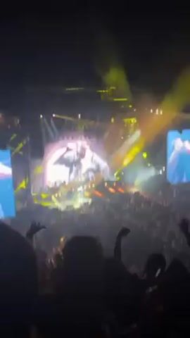 The video captures a live concert with a large audience. The performer is on stage with a band, illuminated by stage lights and projected onto a large screen behind them. The audience is visible in the foreground, with many people raising their hands in the air. There are multiple musicians accompanying the performer. The atmosphere is energetic and filled with smoke effects.