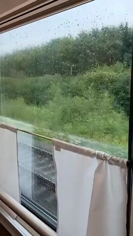The video shows a view from a window, possibly on a train. Rain is falling outside, creating streaks on the glass. Through the rain-streaked window, a blurry green landscape of trees and fields can be seen, moving past as if the viewer is in motion. White curtains hang on the right side of the window frame.