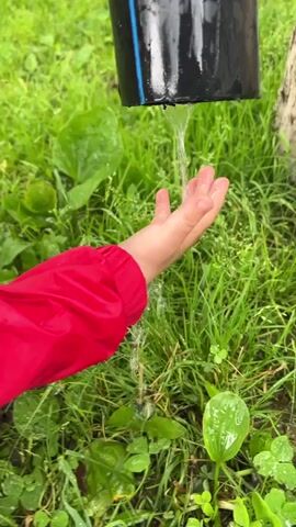 A close-up shot shows a child's left hand wearing a red jacket, catching water from a black pipe. The water streams down from the pipe and splashes onto the child's hand and the surrounding lush green grass. The background is filled with green vegetation and a portion of a tree trunk is visible on the right. The scene appears to be outdoors, possibly in a garden or park. The water stream is continuous throughout the short clip.