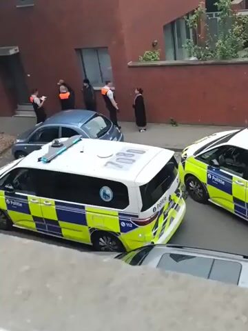 The video shows an outdoor scene with two white police vans, each marked with blue and yellow checkered patterns and the word "POLITIE," parked on a paved street. A smaller blue car is parked behind one of the vans. In the background, several individuals are visible near a reddish-brown building. At least two people are wearing orange safety vests, and others are in darker clothing. They appear to be standing calmly on the sidewalk or street.
