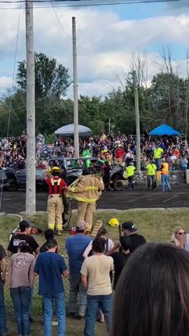A demolition derby event is underway. In the foreground, spectators stand behind a fence, observing the action. In the midground, several individuals in firefighting gear are attending to a severely damaged vehicle, which appears to have rolled over or sustained significant impact. One firefighter is assisting another person, possibly a driver, out of the vehicle. Other emergency personnel and bystanders are also present near the damaged car. In the background, more spectators are seated in bleachers, watching the event. The environment is outdoors, under a partly cloudy sky, with trees and utility poles visible. The atmosphere suggests an active and potentially hazardous event.