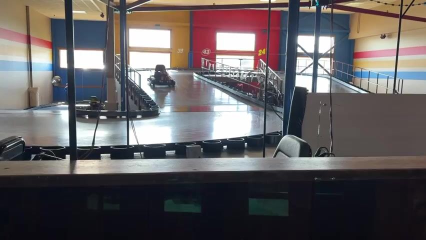 The video shows an indoor go-kart track. In the foreground, a wooden counter is visible with some black chairs behind it. The track itself is made of polished wood, with black tire barriers lining the course. A single go-kart, driven by a person wearing a blue shirt, is navigating a turn on the track. In the background, the walls are painted with colorful stripes, and large windows allow natural light into the building. Another go-kart can be seen in the distance near a ramp. The overall environment suggests a recreational facility.
