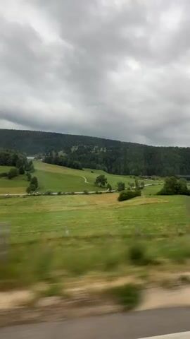 This media file captures a scenic view of a rolling green landscape under a dramatic, overcast sky. The scene is a picturesque rural setting, likely in Petit-Val, Switzerland.

The primary subjects are the natural elements of the landscape. Verdant, well-maintained fields stretch across the frame, punctuated by scattered trees and dense forests that cover the rolling hills in the background. A winding path is visible through one of the fields, suggesting human presence and movement within the landscape. The overall impression is one of serene natural beauty, despite the somewhat moody weather.

The activity taking place is implied through the motion blur of the foreground, indicating that the view is captured from a moving vehicle, likely a car or train. The camera is panning or moving forward, giving a sense of travel through this idyllic countryside.

Notable details include the time of day, which appears to be daytime, possibly late afternoon or early evening, given the soft, diffused light and the dramatic, heavy cloud cover. The weather is clearly overcast, with a thick layer of grey clouds dominating the sky, creating a sense of depth and atmosphere. There are no visible people or specific objects of interest in the frame other than the natural landscape and the implied vehicle. The location cues of rolling hills, dense forests, and well-kept fields strongly suggest a European rural setting, consistent with Petit-Val, Switzerland. The emotion evoked by the scene is one of tranquility and natural beauty, perhaps with a touch of melancholy due to the cloudy sky.

There is no visible text in the media file.