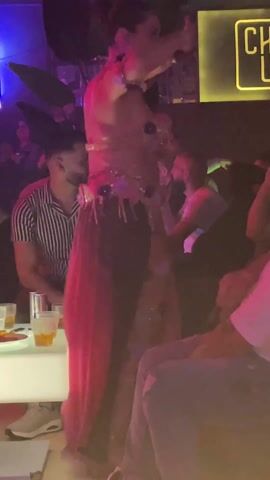 A dimly lit nightclub is the setting for this video. In the foreground, a person adorned in a sheer floral-embellished dress with black and pink panels, and a flower necklace, dances with their back to the camera. A man seated at a table, wearing a vertically striped shirt, attentively watches the dancer. Other people occupy the background. Orange-colored drinks sit on the illuminated table in front of the seated man. The lighting varies between green and blue. A neon sign that reads "Chilai" is partially visible in the upper right corner. Several large artificial plants are placed to the left of the dancer.