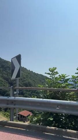 The video begins with a view of a lush green mountain under a clear blue sky. A guardrail runs across the foreground. A small building with a red roof is visible on the mountainside. The perspective shifts to focus on the guardrail and then a dense hedge. The final frames show a brick wall and a closed white door with a black handle.