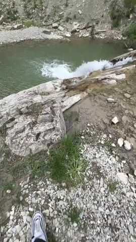 The video depicts a small waterfall cascading into a pool of water. The surrounding environment is rocky with a layered cliff face visible in the background. Some greenery is present, including grass near the camera's position and trees on the hilltop. The water in the pool has a greenish tint, and the waterfall creates a white splash as it enters the pool. The camera seems to be positioned on a rocky outcropping, looking down at the waterfall and pool. The scene is outdoors, and the lighting suggests a partly cloudy or overcast day.