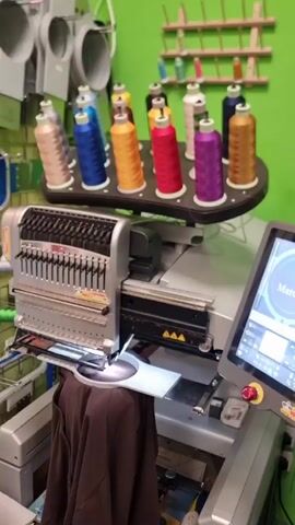 The video shows an industrial embroidery machine with multiple spools of colorful thread mounted above it. The machine has a digital touchscreen display showing the name "Marcela" within a circular design. A person's hand is seen pressing a green button on the control panel of the machine. The background appears to be a workshop or studio with green walls and shelves holding more spools of thread.