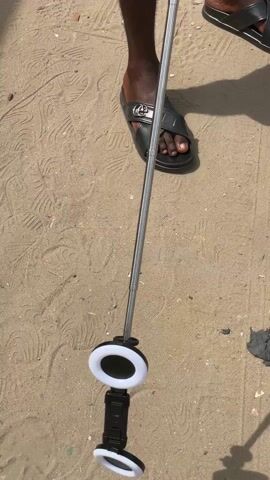 The video starts with a close-up of a person's legs, wearing sandals, standing on sandy ground. The person is holding a selfie stick with a circular light at the bottom. The camera then pans up to reveal a wider scene of a dusty outdoor environment, with a low brick wall and buildings in the background. Laundry hangs on a line attached to the wall. There is trash scattered on the ground. The sky is overcast. The selfie stick is held up in the air.