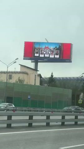 The video shows a billboard from a low angle, as seen from inside a moving vehicle. The billboard features five portraits of people against a background of a red curtain and a cityscape with towers reminiscent of the Kremlin. There are numbers on the sides of the billboard that likely indicate its location. The background environment includes a section of green sound barrier, a building, trees, lampposts, and other vehicles on the road. The sky is overcast.
