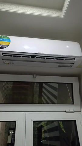 The video shows a close-up view of a white air conditioner unit mounted on a wall above a window. The air conditioner has a digital display on its front panel showing the number 18. There is a circular energy efficiency sticker on the left side of the unit. Below the air conditioner is a white framed window with glass panes. The window frame appears to be part of a larger structure, possibly a cabinet or another set of windows. The ceiling above the air conditioner has decorative molding. The overall scene is indoors, with artificial lighting. The camera is positioned slightly below the air conditioner, looking upwards.