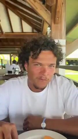 A young man with curly brown hair and blue eyes sits at an outdoor table, looking at the camera. He is wearing a white t-shirt and has a blue bracelet on his left wrist. There is a plate with food in front of him, and he appears to be chewing something. The table is under a wooden structure, possibly a veranda or open-air restaurant, with other people visible in the background at other tables. The setting suggests a relaxed, casual dining environment, possibly on vacation.