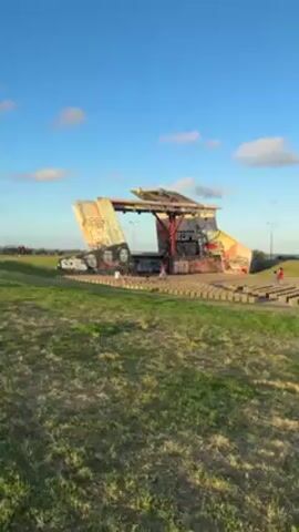 The video shows a graffiti-covered, angular open-air stage with concrete block seating on a grassy area under a partly cloudy blue sky. Several small children are visible, running and playing on the steps and stage. In the distance, some people stand along a horizon line. The structure includes portraits and various graffiti tags, with the phrase "ANTICAYO BI P" visible above an opening. Streetlights stand nearby.