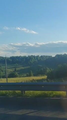 The media file shows a view from a moving vehicle, likely a car, traveling along a road. The scene is a rural landscape bathed in the warm light of late afternoon or early evening. The sky is a vibrant blue with scattered white, fluffy clouds, suggesting fair weather.

The foreground is dominated by a metal guardrail and a concrete barrier, indicating the presence of a road or bridge. Beyond the guardrail, rolling hills covered in lush green trees form a dense forest. Patches of bright green fields are interspersed within the treeline, hinting at agricultural activity or open meadows. A single utility pole stands tall in the middle ground, its dark silhouette contrasting with the bright landscape. Further in the distance, a faint outline of what appears to be a cell tower or transmission mast can be seen atop a hill, a subtle sign of human infrastructure in the otherwise natural setting.

The lighting suggests that the sun is low in the sky, casting long shadows and creating a peaceful, serene atmosphere. There is no visible text in the media file. The overall impression is one of a scenic drive through the Russian countryside, offering a glimpse of nature's beauty under a clear sky.