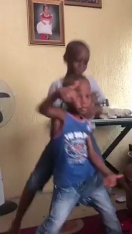 Two young boys are dancing inside a room. The boy in the front is wearing a blue tank top and blue jeans. He has a shaved head. The boy behind him is wearing a gray t-shirt and blue jeans. The walls of the room are a light beige. On the wall behind the boys are two framed pictures. To the right of the boys is a keyboard on a stand, and a fan is visible on the left. The boys are smiling and dancing energetically, moving their arms and bodies to the music.