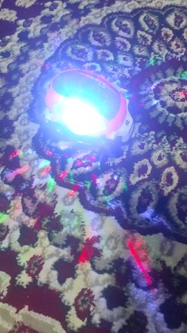 The video shows a toy car with bright lights and a spinning, colorful light show projected onto a patterned carpet. The car is orange and white and seems to be stationary in the center of the frame. The carpet has an organic pattern with dark and light sections. The multi-colored lights from the toy dance across the surface of the carpet.