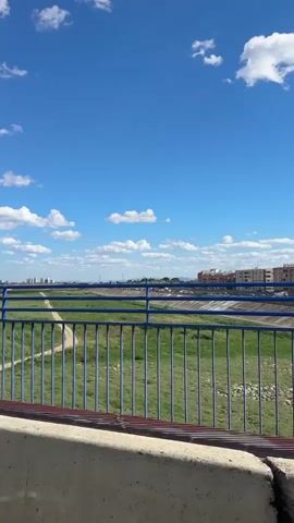The camera is positioned on a bridge, looking out over a riverbed and cityscape on a sunny day with a blue sky and scattered white clouds. A blue metal railing with vertical bars runs along the foreground. Below the bridge, there is a wide, green, grassy area. A dirt path runs alongside what appears to be a concrete river channel. In the distance, there is a row of apartment buildings with various colors. Further in the distance, there is a skyline of buildings and towers under the clear sky. Cars can be seen moving along a road running parallel to the riverbed.