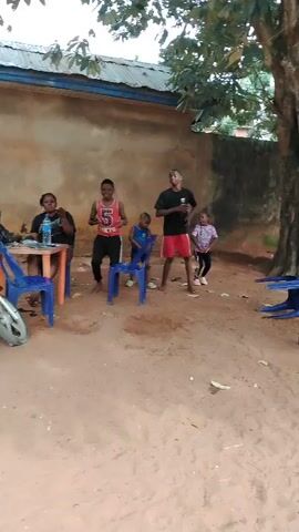 The video shows an outdoor scene in what appears to be a yard or open space with sandy-brown ground, located in Akwa Etiti, Nigeria. A large tree with a thick trunk and foliage is prominently on the right, providing shade. In the background, there's a light-colored building with a corrugated metal roof.

Several people are present, including adults and children. On the left, an adult person is seated at a wooden table. In the center, three children and one adult are standing. A boy wearing a red tank top with "5 TOKYO" printed on it and black shorts is moving, seemingly dancing, next to a smaller boy in a blue shirt and shorts. Another adult male in a black t-shirt and red shorts is also standing and moving with them. A small girl in a patterned top and black bottoms is visible behind this adult. Multiple blue plastic chairs are scattered around the sandy ground, some upright and some overturned. The overall atmosphere seems relaxed and casual, possibly a family gathering or playtime, during daylight hours.