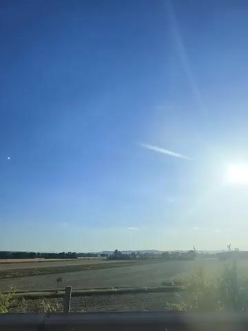 A bright sun shines in a clear blue sky over a rural landscape. In the foreground, the top of a dark horizontal surface is visible, possibly a car dashboard or railing. To the left, a portion of a green leafy tree is in frame. Beyond the foreground, a wide field stretches into the distance, with hints of vegetation and possibly agricultural crops. In the background, a line of trees and structures mark the horizon, with gentle rolling hills visible further back. The overall impression is one of a sunny day in the countryside, possibly viewed from a moving vehicle.