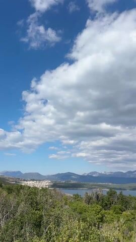 The video presents a scenic view featuring a body of water with mountains in the background, under a sky partially covered with puffy clouds. The landscape in the foreground is lush with green bushes and vegetation. A town can be seen on the shore opposite the viewer. The mountains across the water have snowy peaks. The sky is vibrant blue, peeking through the layers of clouds. A small sailboat is visible on the water.